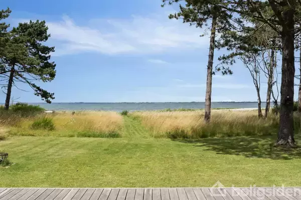 Græsplæne strækker sig mod en rolig kystlinje, flankeret af højt græs og spredte træer, under en klar blå himmel. En træterrasse danner forgrunden.