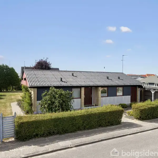 En enkeltstående etplanshus med gråt tag står stille på en stille villavej omgivet af velplejede hække og en græsplæne. Blå himmel ses bagved.