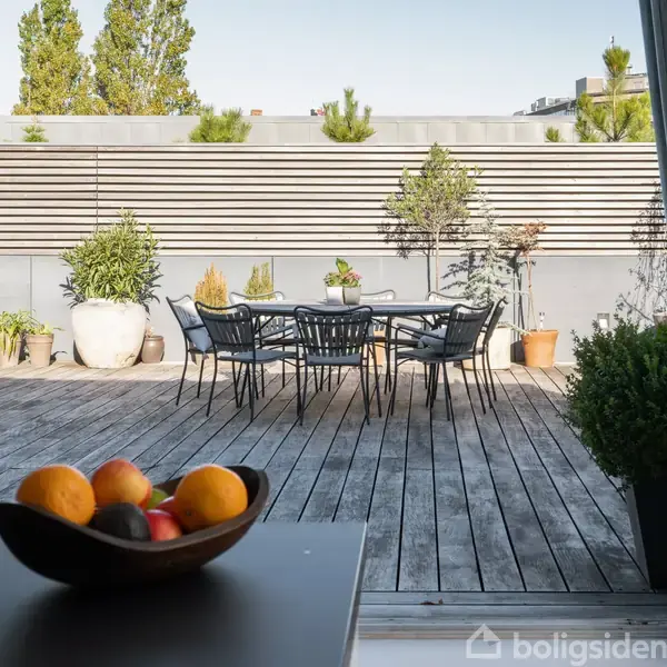 Skål med frugt står på bordet i forgrunden; i baggrunden ses et træterrasseområde med et bord og stole, omgivet af plantepotter foran en skærmvæg med træer.