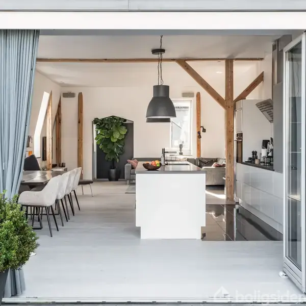 Åben køkken-alrum med hvidt interiør, spisebord med stole til venstre, køkkenø i midten, og planter for enden. Store lamper hænger fra loftet, og rummet er lyst og moderne.