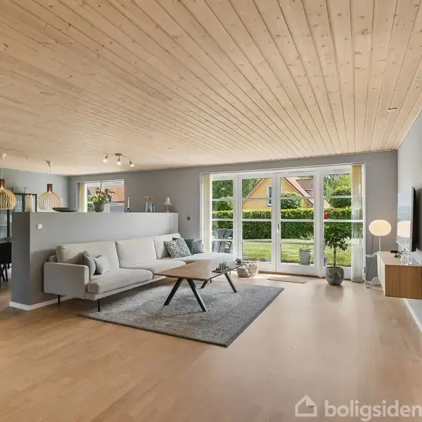 Stue med lyse trægulve og loft med sofa og sofabord på tæppe, stort vindue mod have; spiseplads i baggrunden. Lys og moderne indretning.