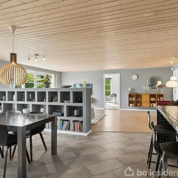 Spisebord med stole står i en moderne stue med trægulv, åbne reoler og store pendellamper. Der er barstole ved en disk og kunst på væggene.