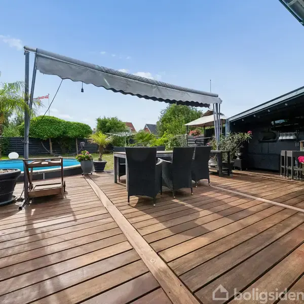Udendørs terrasse med trædæk rummer en spisegruppe under en stor markise. Omgivelserne inkluderer krukker med planter og pool i baggrunden. Højre side viser en pavillon med bar.