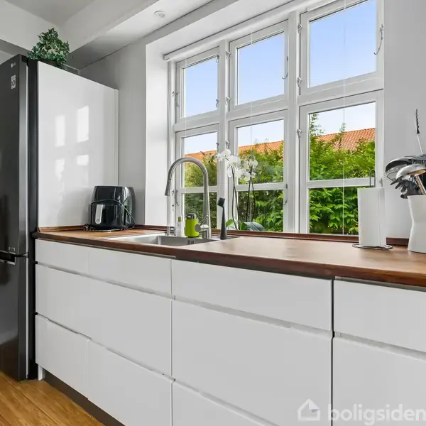 Et moderne køkken med hvide skabe og en sort køleskab. En vask med armatur er centreret under store vinduer, der viser udsigt til en have. En elkedel er placeret på bordpladen.