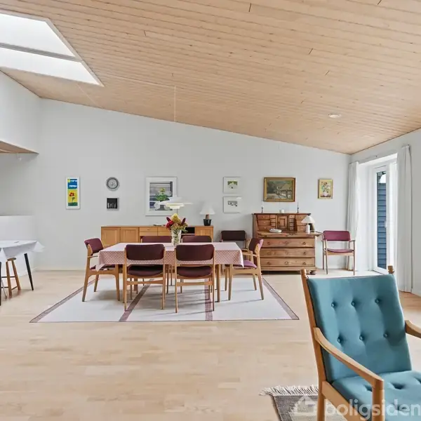 Spisestue med bord og stole centreret på et tæppe, omgivet af billeder og kommoder på væggen. Store vinduer til højre og loftsvindue skaber lys i rummet.
