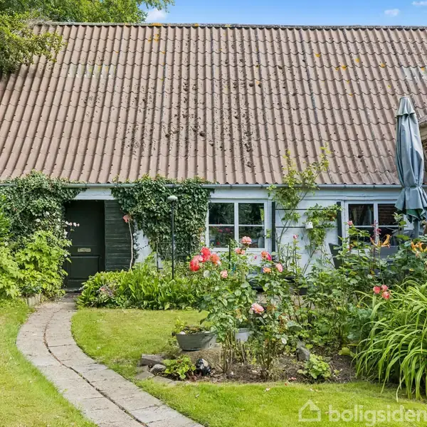 Et hus med et skråt tag dækket af mos. En grøn dør flankeret af vinduer. Foran er en blomstrende have med roser, en stensti og grønt græs.