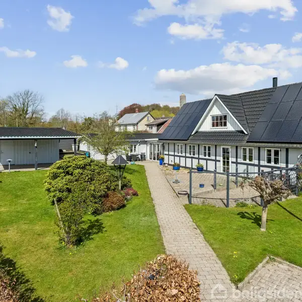 Et hvidt hus med sort tag står i en have med velplejede plæner og en flisebelagt sti i midten. Omkring huset er der buske og et udhus til venstre.