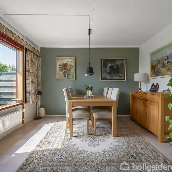 Et spisebord med to stole er placeret på et tæppe i et lyst rum. Bordet har en dekoration. Væggene har malerier, og et stort vindue giver udsigt til haven.