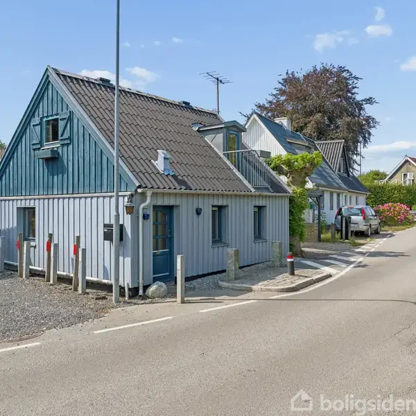 Et blåligt træhus står roligt langs en stille vej i et boligområde. Omgivet af andre huse og grønne træer under en klar blå himmel.
