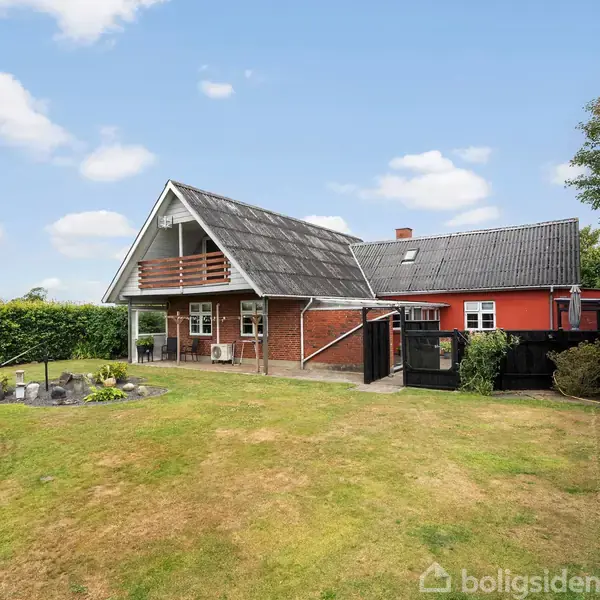 Hus med rødt murværk og sort tag. Terrasse og balkon på forsiden, omgivet af velplejet have med græsplæne og buske. Klar blå himmel med hvide skyer i baggrunden.