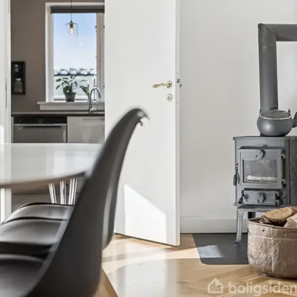 Brændeovn står stille ved væggen i en lys stue; en potte på toppen og brænde i en kurv ved siden af. Åben dør afslører moderne køkken i baggrunden.