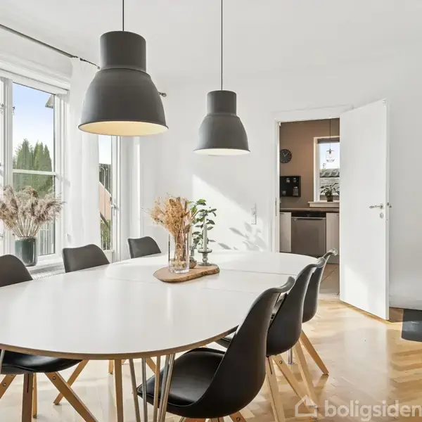 Et ovalt spisebord med sorte stole står i et lyst rum med store vinduer. Over bordet hænger tre sorte lamper. En brændeovn står til højre ved en åben dør.