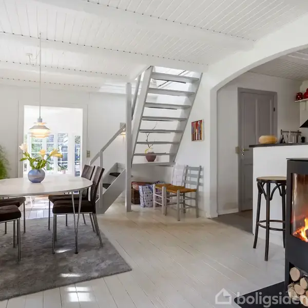 Brændeovn varmer hjørnet af et lyst, moderne rum. Runde spisebord med stole står på gråt tæppe. Trappe fører til ovenpå. Vægge er hvide, og der er kunst på væggene.