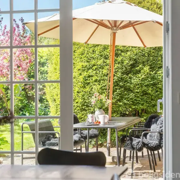 En terrasset, set gennem åbne glasdøre, har sorte stole ved et bord med skygge fra en parasol, mens haver med blomstrende træer og buske udgør baggrunden.