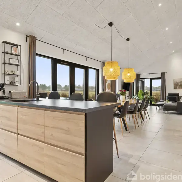 Køkkenø med træskabe står centralt, omgivet af en åben spisestue med sort bord og stole samt opholdsstue. Store vinduer giver udsigt til græsplæne udenfor. Tre lamper hænger fra loftet.