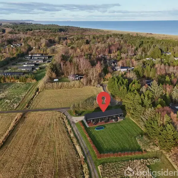 Et hus markeret med en rød placemarkør er omgivet af træer og marker, med en vej foran. I baggrunden ses der skov og en kystlinje.