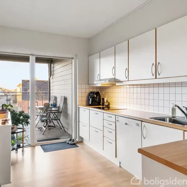 Et køkken med hvide skabe og træbordplader, hvor en kaffemaskine står på bordet. Gennem en glasskydedør ses en altan med et lille bord og to stole.