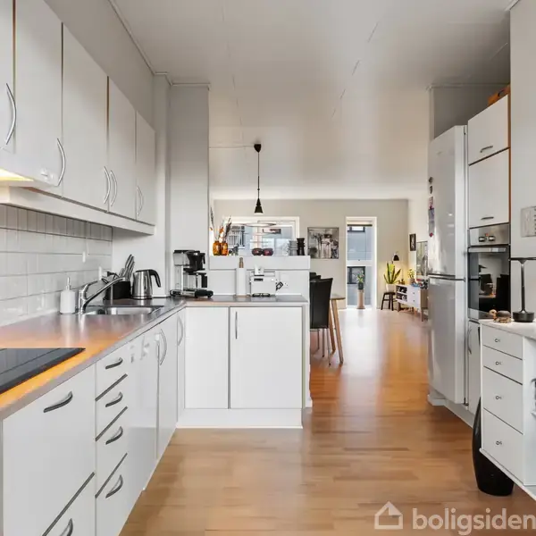 Et lyst køkken med hvide skabe og skuffefronter, en induktionskogeplade og en hvid flisevæg. Køkkenet fører ind til en spisestue med træstole og diverse dekorationer på væggene.