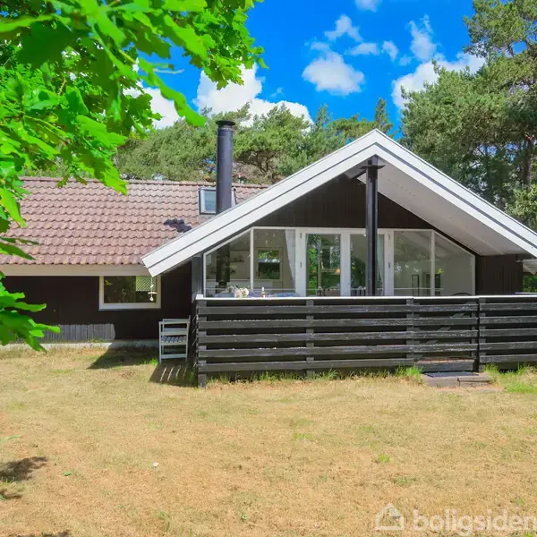 Sommerhus med sort træfacade og terrasse står i sollys. Omgivet af græsplæne og træer, under en klar blå himmel med skyer.