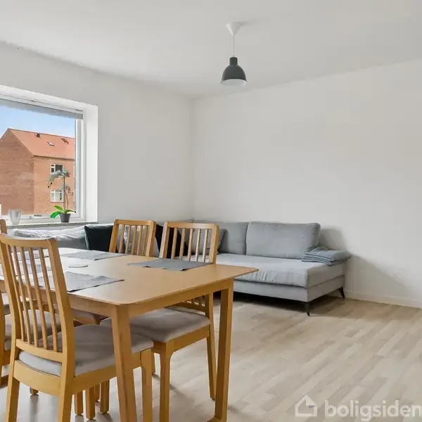 Et lysebrunt spisebord med seks stole står på et lyst parketgulv i en stue. Sofa ved hjørnet, glasvitrineskab til venstre, og en lampe hænger fra loftet. Et vindue med udsigt til en rød murstensbygning.