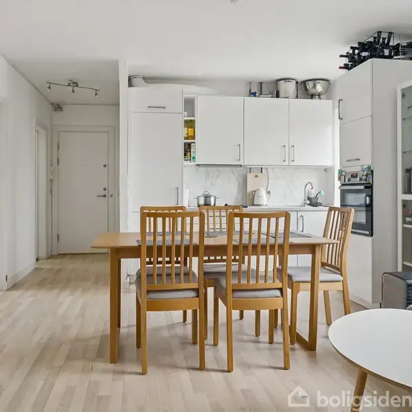 Et spisebord med seks stole står centralt i et lyst køkken-alrum, hvor der er hvide skabe, en ovn og en glasvitrineskab. Lejligheden har lyst trægulv og minimalistisk indretning.