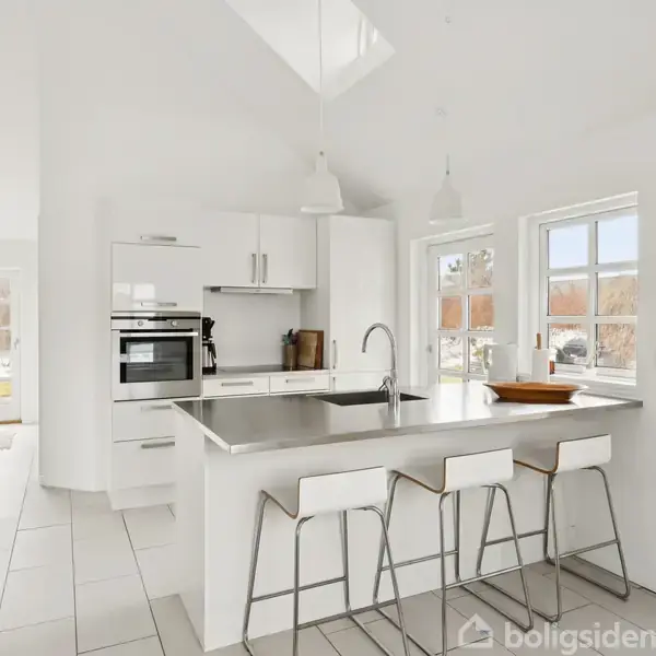 Hvidt moderne køkken med køkkenø og tre barstole. Køkkenet har indbygget ovn og vinduer, der viser en have udenfor. Lyse fliser på gulvet og minimalistisk indretning.