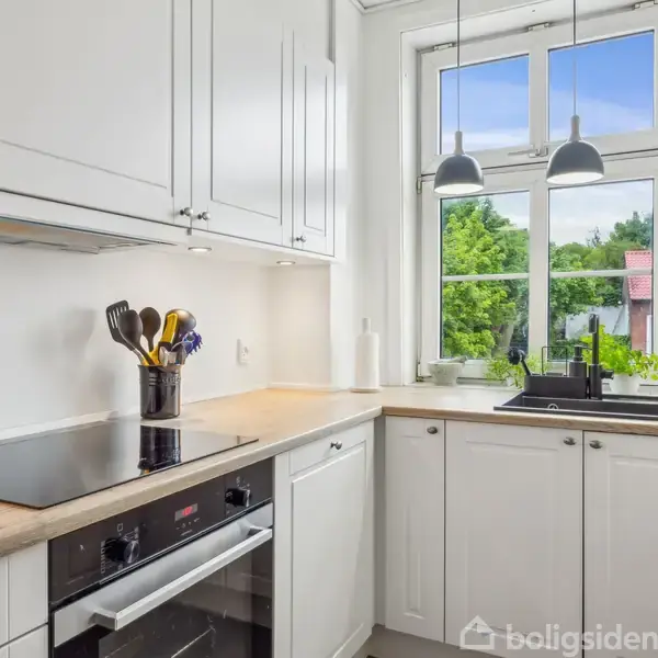 Et lyst, moderne køkken med hvide skabe, indbygget ovn og kogeplade. Køkkenredskaber står i en beholder. Et vindue med udsigt til træer giver naturligt lys, flankeret af loftlamper.
