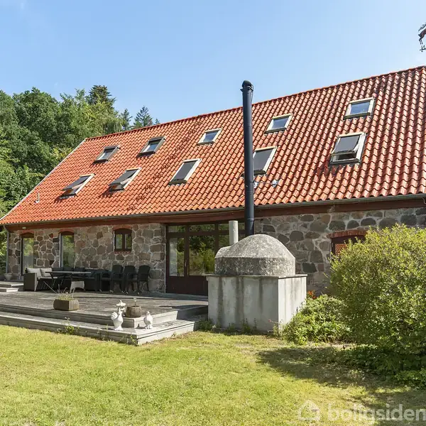 Stenhus med rødt tegltag står i en frodig have omgivet af skov. Flere tagvinduer ses, og en terrasse med havemøbler er foran huset.