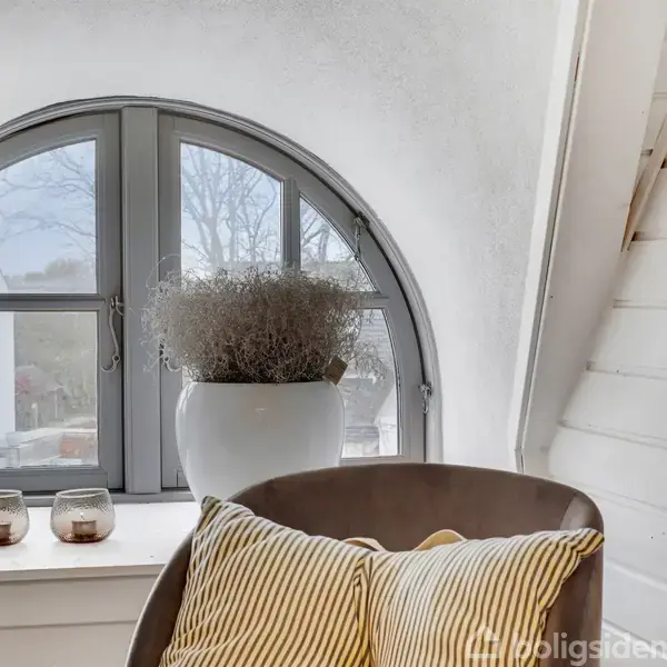 En stol med stribede puder står foran et buet vindue med klart glas. På vindueskarmen er der en plante i en hvid potte og to små stearinlys holdere.