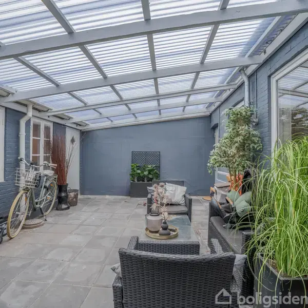 Et overdækket terrasseområde med rattanmøbler og grønne planter, omgivet af blå murstensvægge. En cykel står til venstre, og et gennemsigtigt tag lukker lys ind.