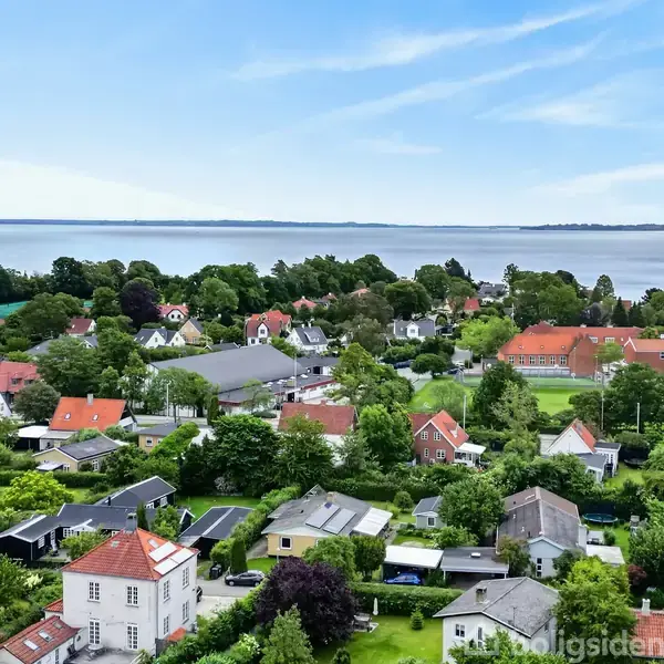 Række af huse står stille blandt grønne træer; skaber et behageligt boligkvarter. I baggrunden ses en rolig kystlinje med havudsigt under en blå himmel.