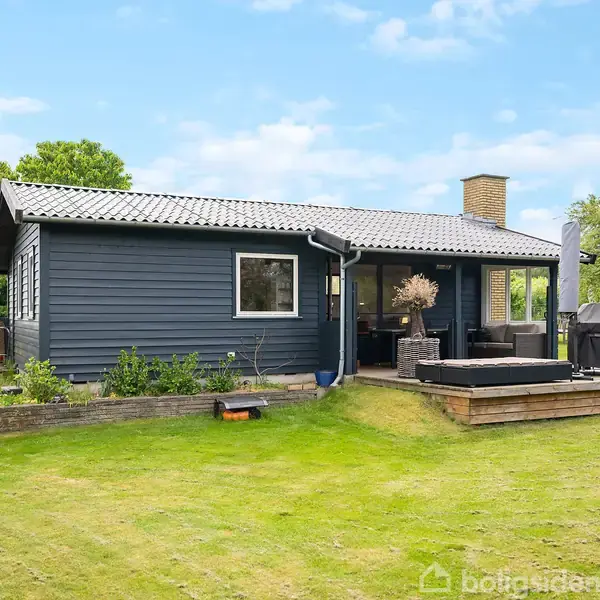 Blåt træhus med terrasse; står stille på en velholdt græsplæne omgivet af træer og buske; terrassen har havemøbler og grill; lyseblå himmel uden skyer.
