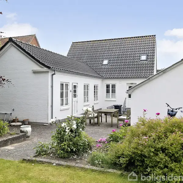 Et hvidt murstenshus med mørkt tag står ved en brostensbelagt terrasse. Et træbord med stole er placeret udendørs, omgivet af blomsterbede og en græsplæne i en have.