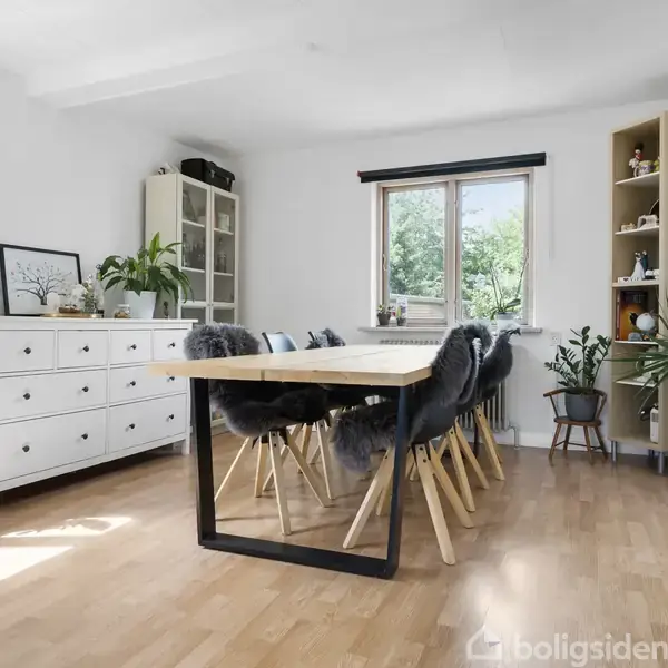 Spisebord med grå polstrede stole placeret i et lyst rum. Omgivet af hvide kommoder, planter og hylder ved vinduet med udsigt til grønne træer.