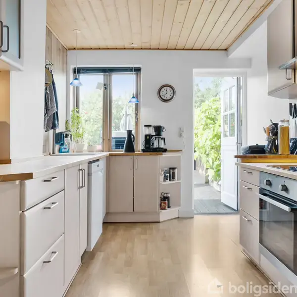 Køkken med hvide skabe og træloft, hvor en kaffemaskine står på bordpladen. Ovn og køkkenmaskine er på hver side, med udsigt til en lysfyldt patio gennem åbent dørparti.