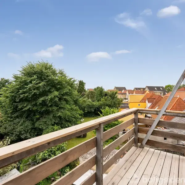 Balkon med trægelænder åbner ud mod en rolig udsigt over hustage og frodige træer, under en blå himmel med lette skyer. En åben dør fører ind til bygningen.