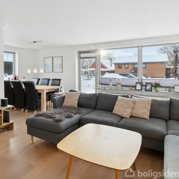 Stor, grå sofa med puder står foran stort vindue. Inde i stuen er der et træbord med sorte stole. Udendørs ses snedækket landskab.