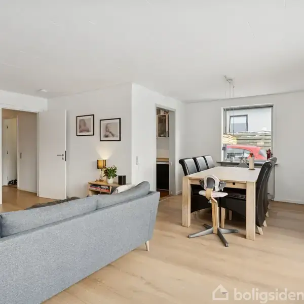 Grå sofa står i en moderne, lys stue med trægulv, vendt mod et fjernsyn. I baggrunden er spisebord med sorte stole foran et vindue, omgivet af neutral indretning.