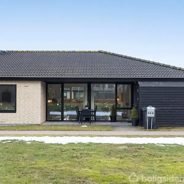 En etplans villa med lys murstensfacade og sorte trædetaljer har en terrasse med havemøbler foran. Træ og buske omkranser huset. Skiltet ved indkørslen viser "44A".