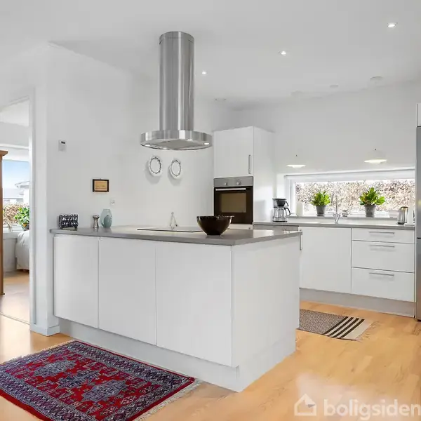 Køkken med moderne køkkenø, hvor en vase og en skål står. Lys indretning med ovn og køleskab. Trægulve og tæpper. Planter i vinduet skaber en frisk atmosfære.