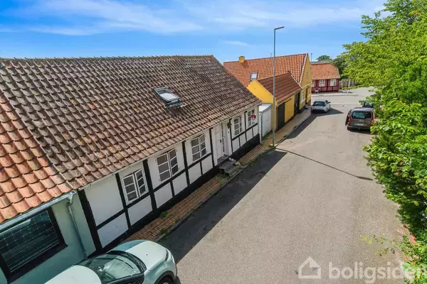 Traditionelt bindingsværkshus med tegltag står stille langs en stille vej. Flere biler er parkerede tæt ved huse med farverige facader. Solen skinner, omgivet af grønne træer.