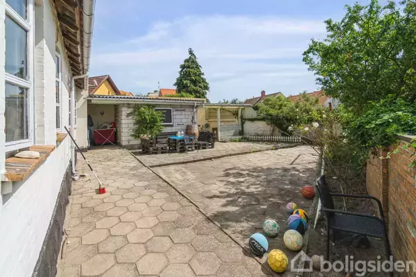 En terrasse med fliser har flere farverige bolde spredt langs en bænk. I baggrunden står et bord med stole ved siden af en hvid bygning. Træer og buske omslutter området.