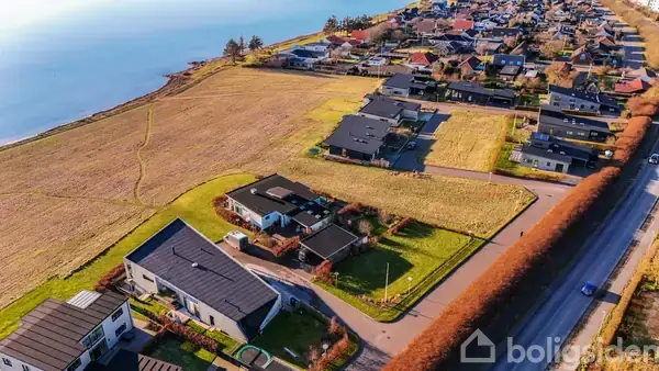 Flere moderne huse ligger langs en vej med hække, tæt på en kystlinje og store åbne marker. Husene er spredt ud og omgivet af græsplæner, med vandet synligt i baggrunden.