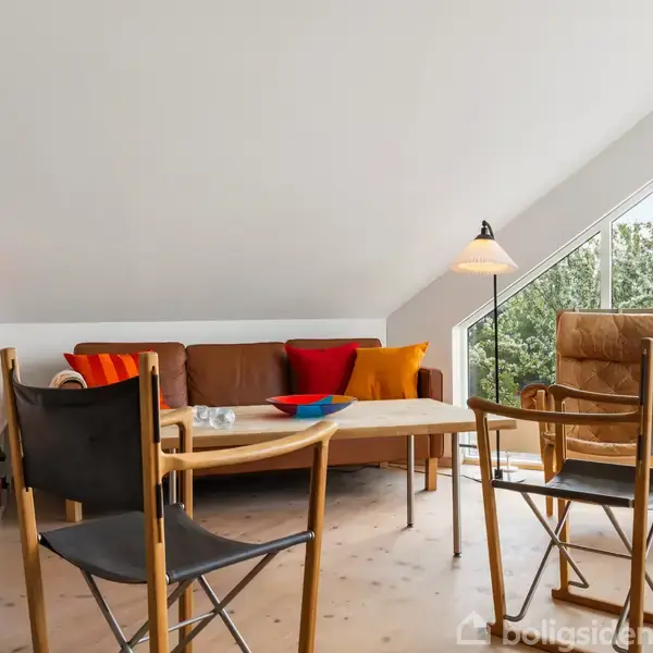 Sofa med farverige puder står i en stue med skrå loft. To stole ved et træbord, en gulvlampe ved stort vindue og hylder med bøger.