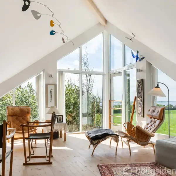 Et lyst stuerum med højt til loftet har moderne møbler og store vinduer, der afslører en udsigt over en grøn mark og blå himmel. Kunstværker hænger fra loftet.