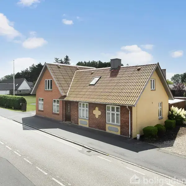 Et gult hus med rødt tegltag står ved en vej. Der er vinduer med hvide rammer, og huset er omgivet af en grønt område med buske. Klare vejrbetingelser viser lys blå himmel.