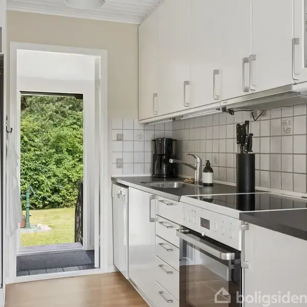 Et hvidt køkken med integrerede skabe og skuffer, sort bordplade og moderne apparater. En dør i rummet åbner ud til en frodig have med grønne træer og buske.