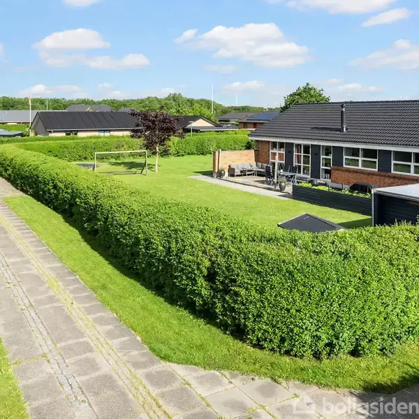 En rød murstensbungalow med sort tag står i et beboelsesområde. Ved siden af huset er en garage, og der er en asfaltindkørsel omgivet af velplejede grønne hække og en græsplæne.