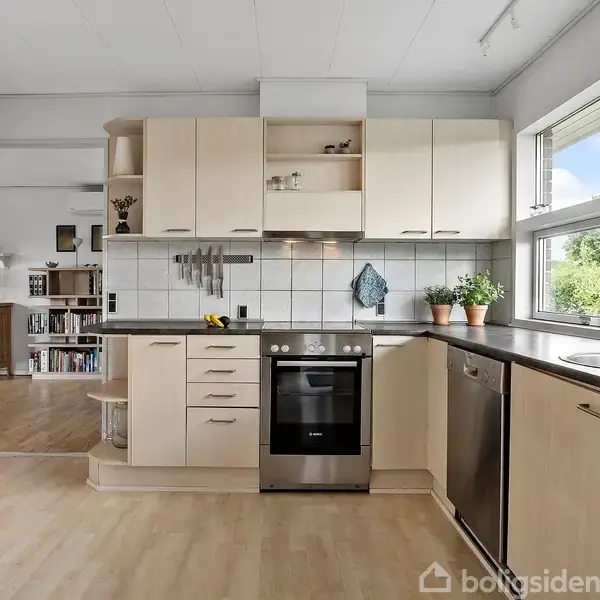 Køkken med træskabe og sort bordplade, moderne komfur og ovn i midten. Rummet har store vinduer med planter på vindueskarmen. Til venstre ses delvist et tilstødende rum.