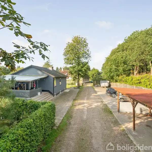 Et gråblåt hus med overdækket terrasse står ved en grusvej, omgivet af grønne hække og skov. På højre side ses en carport med parkerede biler.